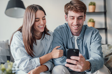 Happy millennial couple sit relax on couch in living room watching video on cellphone together,...