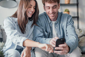 Happy millennial couple sit relax on couch in living room watching video on cellphone together,...