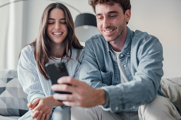 Cute couple greeting having fun video calling long distance friends and family catching up. Young man holding phone with girlfriend sitting on the sofa laughing and enjoying technology