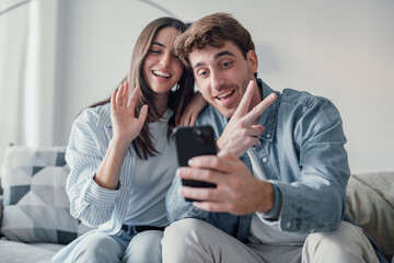 Fototapeta premium Cute couple greeting having fun video calling long distance friends and family catching up. Young man holding phone with girlfriend sitting on the sofa laughing and enjoying technology