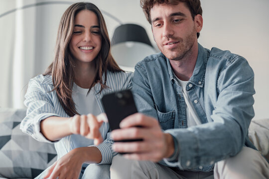 Happy millennial couple sit relax on couch in living room watching video on cellphone together, smiling young husband and wife rest on sofa at home browsing Internet using modern phone device