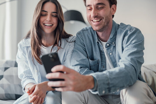 Happy millennial couple sit relax on couch in living room watching video on cellphone together, smiling young husband and wife rest on sofa at home browsing Internet using modern phone device