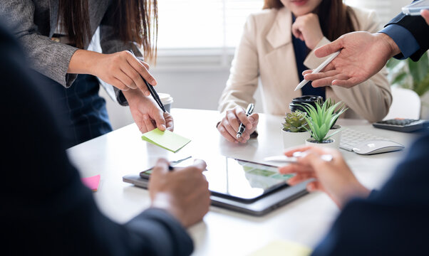 Colleagues are discussing ideas using digital tablet, sticky notes, and pens during a brainstorming meeting around a desk with potted plants and technology tools.