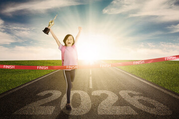 Young girl crossing the finish line and holding a trophy with the year 2026 on the road.