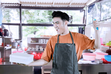 Happy waiter or barista celebrating while holding a takeaway box and a whisk, with a cafe kitchen...