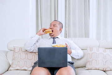 Remote businessman choosing to eat a hamburger while working on a laptop at home