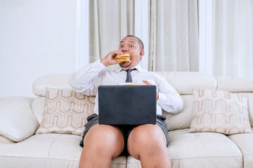 Remote businessman eagerly eating a hamburger while working on a laptop at home