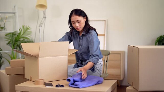 Asian woman packing moving boxes clothes preparing for home relocation apartment change decluttering organizing household items new house setup