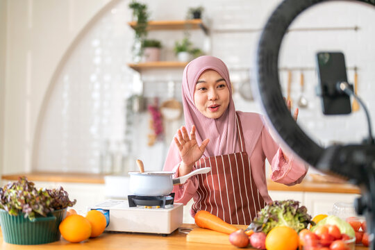 Muslim woman vlogger live streaming cooking online in kitchen look at camera with vegetables, content creator culinary class at home studio, halal food influencer cooking live, video for social media - Powered by Adobe