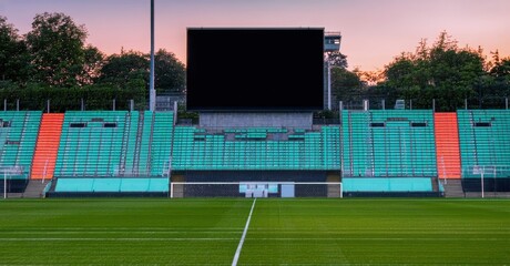 Fototapeta premium Empty stadium view. Green field, colorful seats, large screen visible. Sunset hues paint sky, creating calm atmosphere.
