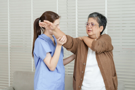 Asian female physiotherapist helps senior asian woman lift arms sideways for shoulder rehabilitation exercise with careful hand support during physical therapy session in indoor clinic