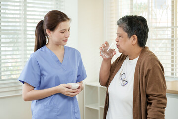 Senior asian woman drinking water after taking medicine with young asian female physiotherapist...