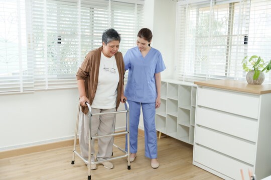 Senior asian woman using walker during home rehabilitation session guided by young asian female physiotherapist in uniform walking beside offering gentle support for elderly physical recovery