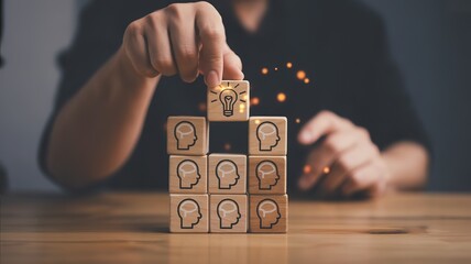 Person stacking wooden blocks with head and lightbulb icons, symbolizing idea generation and teamwork