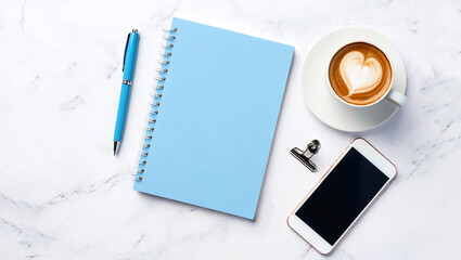A high-angle, flat lay view of a light blue spiral-bound notebook, a cup of latte with a heart foam design, a smartphone, and a pen on a white marble surface.