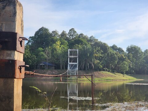 Fototapeta Old rope bridge over calm pond leading to forest observation tower with tropical trees in background. Peaceful rural scene under blue sky.