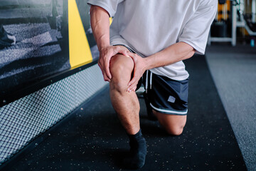 A close-up shot of a man holding his knee in pain, kneeling on the gym floor. The image focuses on...
