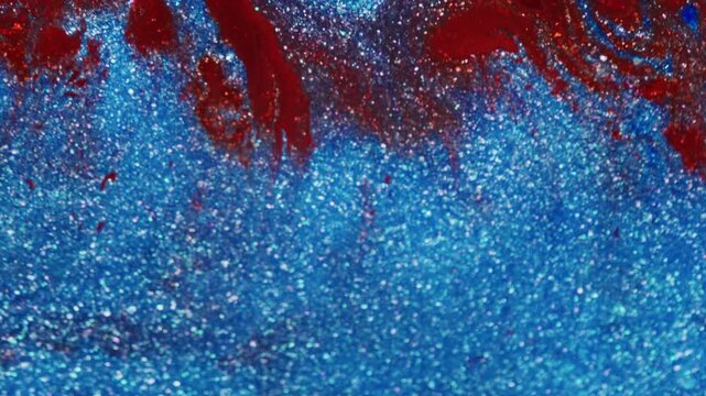 Macro shot showing red and glittery red ink being pushed into a shimmering blue glitter ink surface with visible liquid motion.