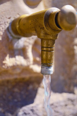 Close-up of an old brass faucet pouring fresh water into a stone fountain, providing a source of...