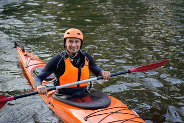 Smiling man paddles confidently in a bright orange kayak, training on a tranquil river surrounded...