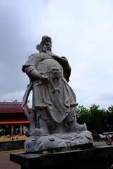 Fototapeta premium Low-angle view of a carved stone guardian statue standing tall at Sam Poo Kong temple in Semarang, Indonesia, under cloudy skies.