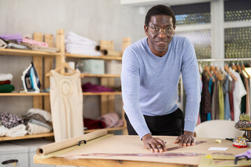 Thoughtful experienced male dressmaker sketching clothing pattern manually, carefully shaping outlines for custom piece on paper, in sewing workshop