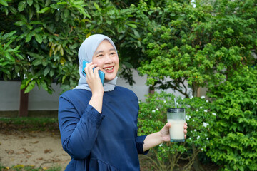 Asian hijab woman holding reusable glass cup with iced milk drink, talking on phone in green garden. Concept of eco-friendly, zero waste, and plastic-free lifestyle.