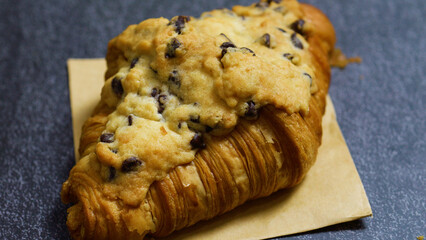 Freshly baked crookie croissant topped with a chocolate chip cookie, resting on brown paper against a dark background, creating a tempting pastry combination