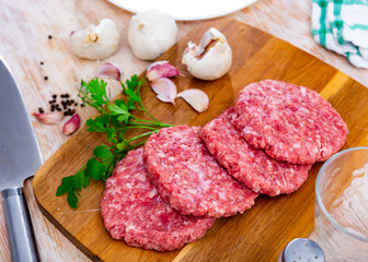 Raw formed hamburger patties ready for cooking on wooden cutting board with fresh fragrant parsley, spicy garlic and allspice