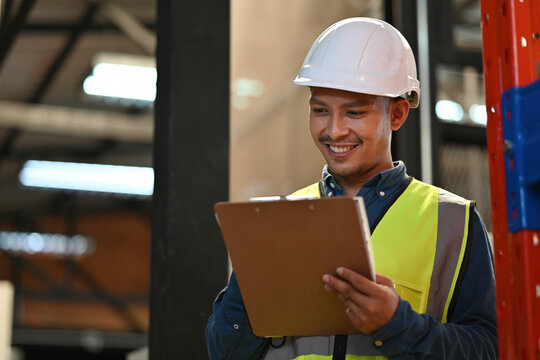 An Asian male worker wearing safety gear writing on a clipboard while inspecting stock in a warehouse - Powered by Adobe