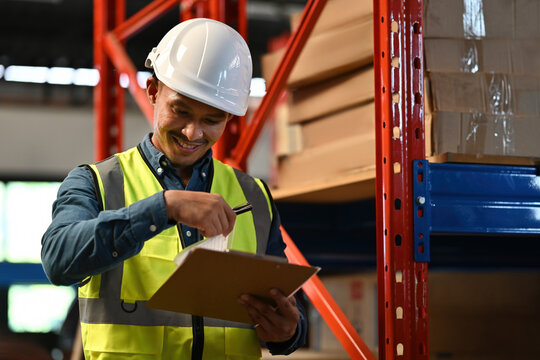 A male warehouse worker wearing a safety vest and hard hat smiles while holding a clipboard and checking stock, symbolizing accurate inventory management and positive work environment