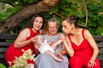 Three generations of women tenderly engage with a baby outdoors on a garden bench, sharing smiles,...