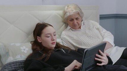 Intergenerational moment capturing grandmother receiving digital explanation from young granddaughter while relaxing together on comfortable bed, sharing technology and family connection - Powered by Adobe