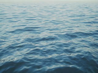 Calm, blue ocean water with gentle, rolling waves under a pale sky.  The surface displays subtle textural variations in light and shadow
