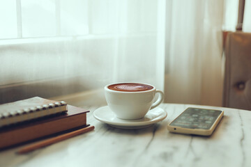 white cup of coffee with latte coffee and smartphone, notebook, pencil on table by the window in...