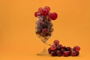 clear glass filled with red and purple grapes stands on an orange background, with a small grape cluster resting beside the base, creating a vibrant and elegant contrast