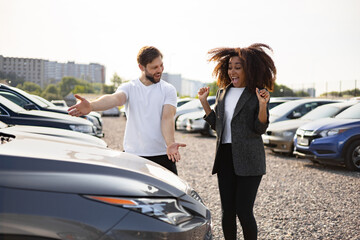 A man showing a car to a woman, who looks excited. They are at a car dealership on a sunny day.