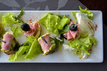 Served on white plate, refreshing salad features crispy lettuce and arugula topped with slices of ham, cucumbers, and olives, creating vibrant restaurant presentation perfect for lunch or dinner.