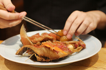 A person's hands use chopsticks to pick at a portion of a braised or stewed fish dish served on a simple white plate
