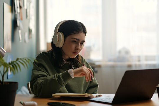 Caucasian teenage girl wearing headphones studying at desk, looking at laptop screen and taking notes in notebook, focused expression, sitting in modern home interior