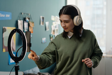 Young adult Caucasian woman wearing headphones interacting with smartphone mounted on ring light tripod in bedroom, recording video content or livestreaming for social media platform