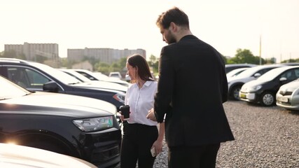 A male car dealer hands over keys to a smiling woman in a car lot, likely after a purchase.