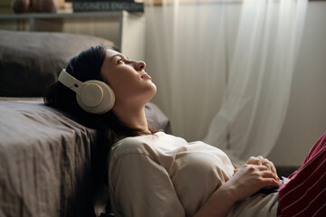 Caucasian young woman lying on floor beside bed listening to music with over ear headphones, gazing...