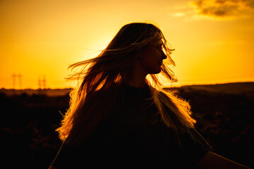 Silhouette of a young woman with long hair blowing in the wind, standing outdoors during sunset...