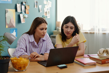 Two Caucasian teenage girls sitting at desk using laptop together, focusing on screen, studying or working on project in home environment with notebooks and snacks visible