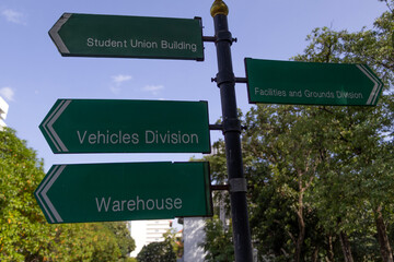 A bilingual direction signpost located inside a Thai university campus, showing directions to buildings in both Thai and English, surrounded by nature and modern architecture.