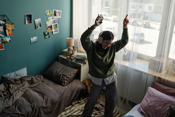 Teenage Caucasian girl standing in bedroom dancing with headphones on, holding smartphone in one hand, smiling with eyes closed, surrounded by personal items and photos