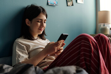 Caucasian teenage girl sitting on bed using smartphone, leaning against wall with relaxed expression, holding device in both hands, engaging with screen content