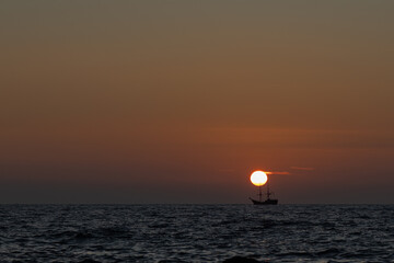  A silhouette of a sailing ship on the horizon during sunset, with the sun perfectly aligned above its mast over calm sea waters.