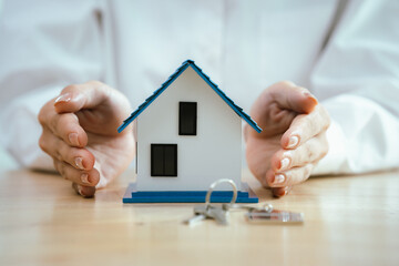 Hands carefully hold and shield a small house model, symbolizing protection, home insurance,...